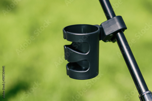 Close-up of black stroller cup holder mounted on handlebar against blurred green park background, minimal modern accessory for parents, outdoor lifestyle and product advertising concept