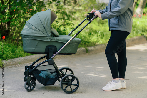 Young woman pushing stylish baby stroller along quiet park alley, mid shot with soft greenery and sunlight, relaxed parenting routine and modern urban family lifestyle concept for advertising