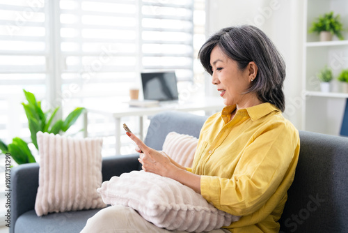 Happy middle aged asian woman using smartphone sitting on couch at home. holding cellphone browsing internet, texting messages on mobile cell phone technology.