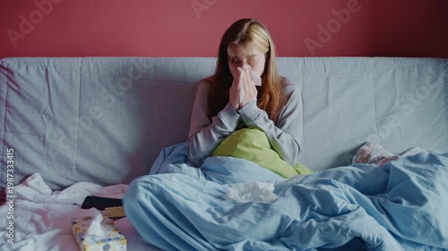 A young woman with the flu lies in bed, feeling unwell as she sneezes into a tissue. Medium shot