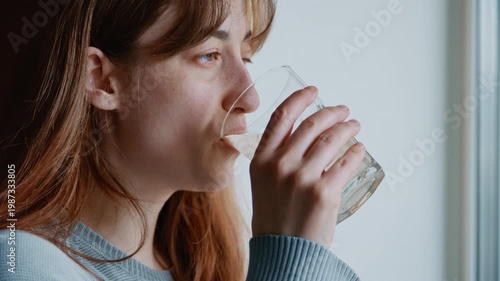 Young woman feeling unwell drinking a glass of medicine while looking out the window