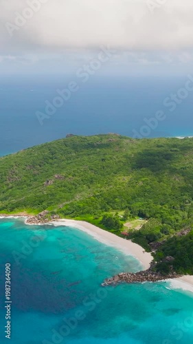 Turquoise ocean waves hitting a rocky shoreline bordered by green forested hills. La Digue, Seychelles.