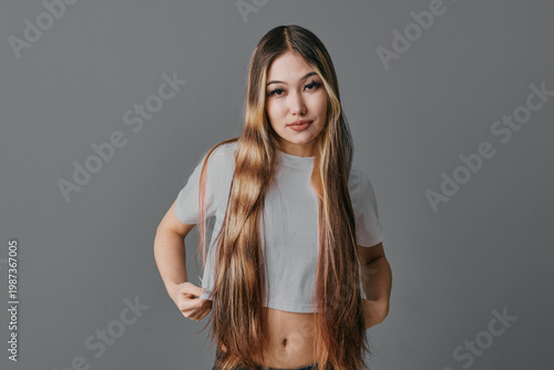 Fotografi Young woman with long straight hair wearing cropped t shirt looks at camera with confident expression on gray background