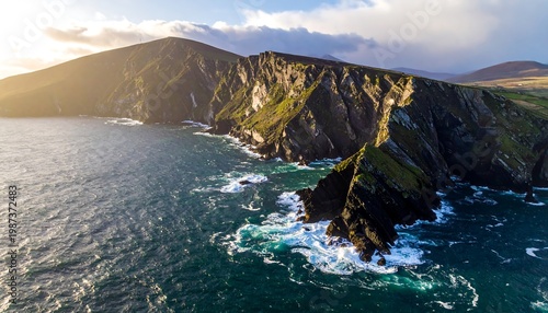 An aerial vista showcases a rugged coastline, where vibrant green cliffs meet a tumultuous ocean under a golden sunlight. Clouds linger in the distance