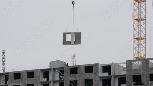 Hoisted wall panel lowered toward rooftop, worker signaling from roof edge, careful alignment with slot, tensioned cables, cloudy day