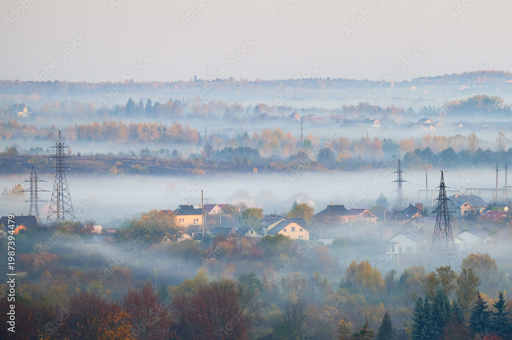 Fototapeta premium Fog drifts over suburban houses and autumn forest in layered morning landscape with power lines. Suitable for weather, environment, energy infrastructure, rural living or seasonal themes