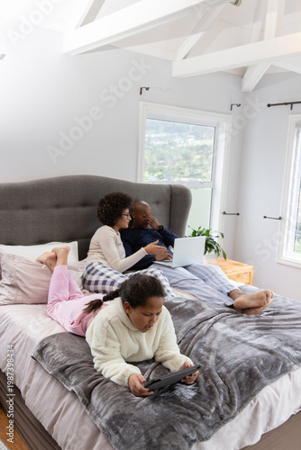 African American family reclining in pajamas on bed, parents sharing laptop, child using tablet