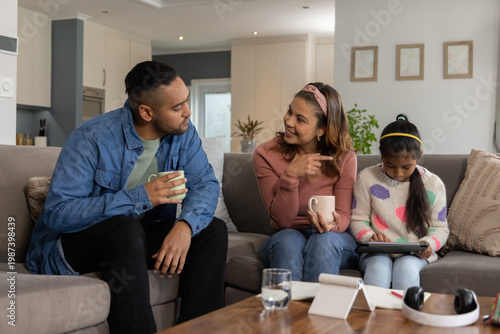 Family sitting on gray sectional sofa at home, parents holding mugs, child using tablet