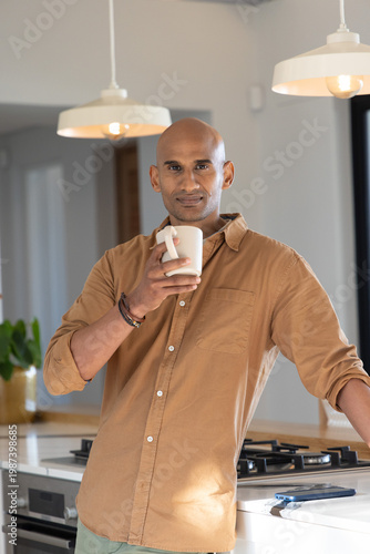 Indian man in 30s sipping from white ceramic mug at kitchen counter with stovetop and plant