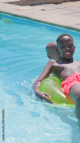 Vertical video: Helping dad steadying son in red trunks boarding green tube at pool, learning swim