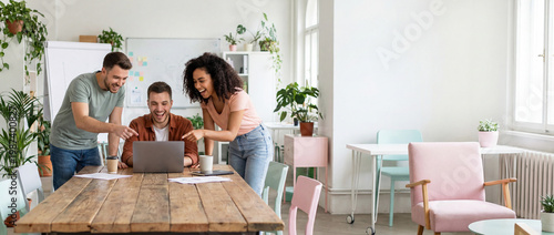 Three happy diverse business colleagues laughing and pointing at a laptop together at a wooden desk in a modern office