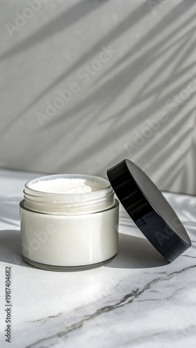 Open jar of white cream with black lid on marble surface with leaf shadows