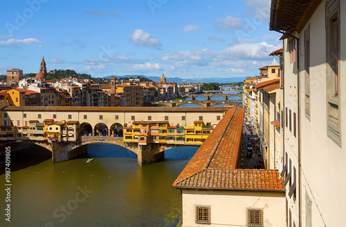 Elevated view of Ponte Vecchio spanning the Arno river in Florence, Italy, with historic architecture, warm rooftops, and bright blue sky above the old city.