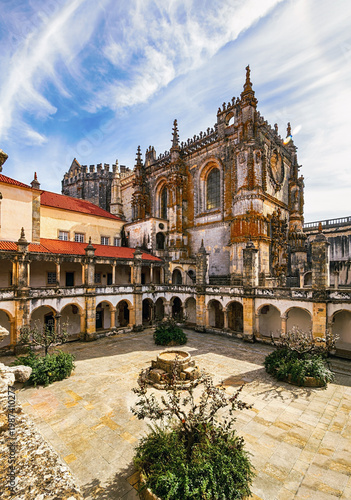 Vertical view of Convent of the Order of Christ (Convento de Cristo), Tomar, Portugal courtyard featuring ornate church architecture, stone arcades, weathered walls, and dramatic clouds in warm daylig
