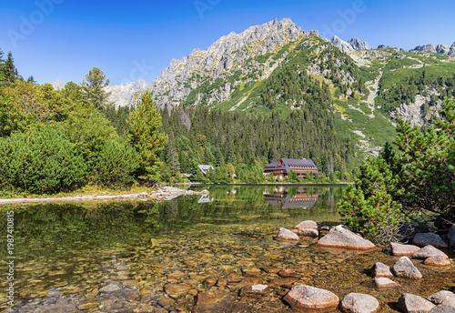 Clear view of Popradske pleso in Slovakia, with rocky alpine mountains, evergreen forest, calm water, and bright summer light in a tranquil natural landscape, Slovakia.