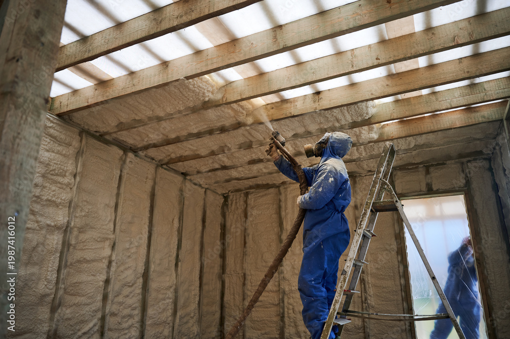 Fototapeta premium Installer covering overhead framing with spray foam insulation in house under construction. Ladder access, protective suit and energy saving building envelope concept