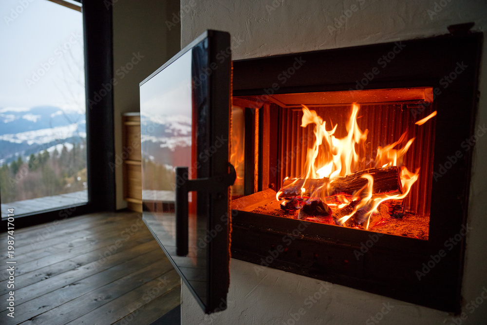 Obraz premium Fire burns in modern fireplace inside cozy home. Close up of glowing logs and flames, glass door open, wooden floor and mountain view through window, warmth and comfort concept