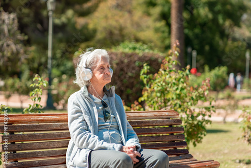 An 85 year old grandmother enjoys music with headphones while sitting on a park bench