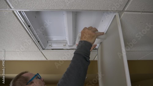 Old middle aged man changing a u-shaped light bulb in a ceiling light fixture in a drop ceiling in a basement