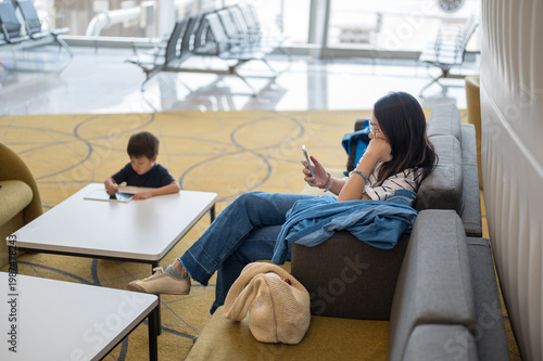 Woman Using Smartphone While Child Engages with Tablet in Modern Airport Lounge