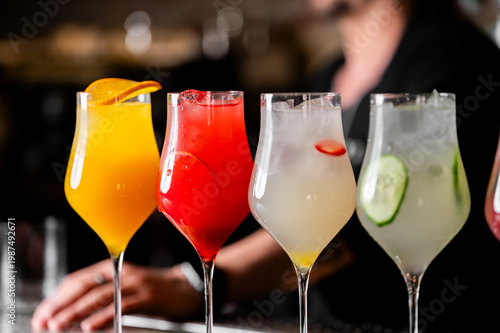 Four vibrant, colorful mocktails or cocktails in elegant tulip glasses lined up on a bar counter, garnished with orange, strawberry, and cucumber. A bartender is blurred in the background.