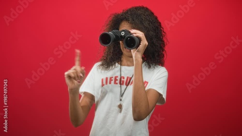 Woman lifeguard in lifeguard tshirt holds binoculars to her eyes and points index finger against a red studio backdrop; alert duty.