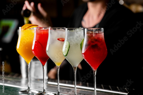 Five vibrant cocktails in elegant tulip glasses lined up on a bar, with a bartender blurred in the background. Each drink features a unique color and garnish like orange, cucumber, and chili.
