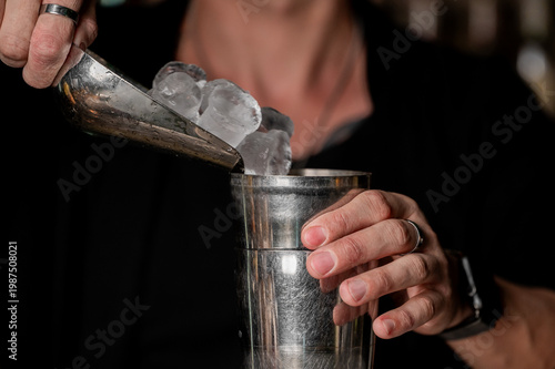Close-up of a bartender's hands pouring fresh ice cubes from a metal scoop into a stainless steel cocktail shaker, preparing a drink at a bar.