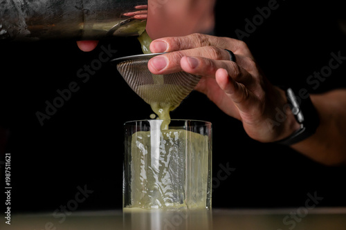 Close-up of a bartender double-straining a green cocktail from a metal shaker through a fine mesh strainer into a glass with a large clear ice cube. Dark background