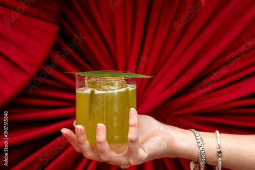 A hand holds a vibrant green cocktail in a rocks glass with a large ice cube and leaf garnish, set against a striking red pleated fabric background.