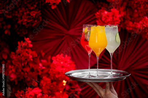Three elegant cocktails—red, orange, and pale yellow—are served on a silver tray against a vibrant red floral and fan background. Perfect for luxury events and celebrations