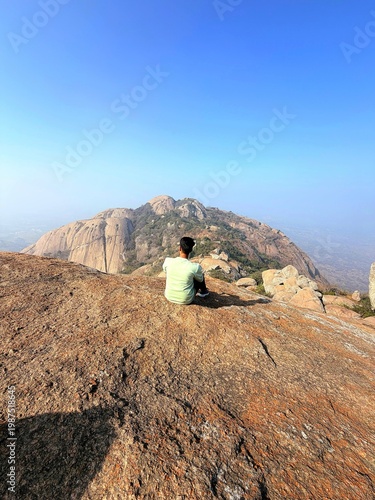 A person sitting on hilltop rock with clear sky and mountain peaks in background, shot from behind