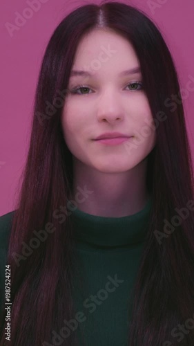 Portrait: young smiling Caucasian woman teen with long naturally black hair. White teenager brunette pretty girl, with green pullover smiles warmly at the camera. Pink background