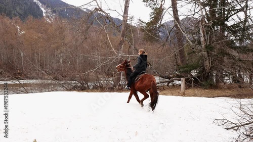 A girl rides a horse in the snow against the backdrop of a mountain river and snow-capped mountains.