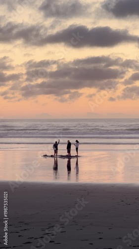 KAWHIA, NEW ZEALAND., 7 April 2026 Three people stand on a Kawhia beach at sunset, looking out at the ocean as waves roll in. The sky is filled with dramatic clouds in shades of orange and gray.