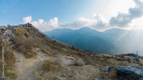 Tableau sur toile View of the Dalai Lama Hills near Mussoorie, Uttarakhand, with colorful prayer flags, sunlit hills, mountain layers in the background, and sun rays breaking through clouds on a winter evening