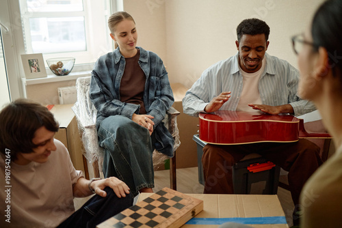 Valokuva Multiethnic young adults socializing during home gathering, playing guitar and board game while unpacking