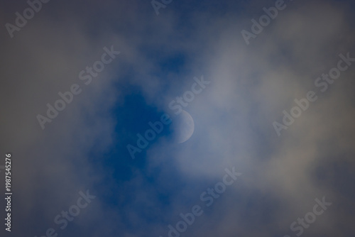 Crescent moon peeks through soft, drifting clouds. Dark blue sky behind. Clouds partially obscure moon. Serene night sky scene. Minimalist composition, peaceful atmosphere