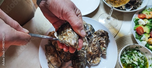 A close-up of hands opening fresh oysters with a knife over a plate. Concepts of fine dining, seafood delicacies, restaurant dining, home cooking, nutrition, and authentic gastronomic experiences.