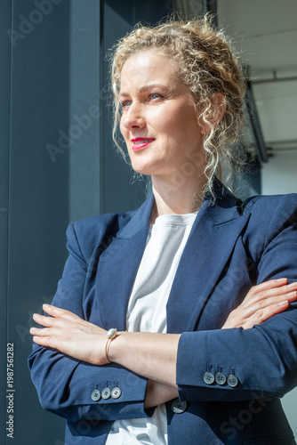 Business woman looking through window with arms crossed in blue blazer, thoughtful confident mood, sunny day corporate lifestyle with copy space