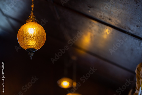 Warm, glowing pendant light hangs from dark wooden ceiling. Soft golden light creates cozy atmosphere. Blurry background enhances depth of scene. Faint reflection visible on ceiling surface