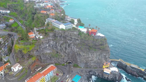 Aerial view of Ponta do Sol, Madeira. Colorful buildings cling to cliffs overlooking the Atlantic, with a charming fishing village and dramatic coastline under a cloudy sky.