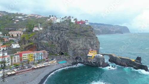 Aerial view of Ponta do Sol, Madeira. Colorful buildings cling to cliffs overlooking the Atlantic, with a charming fishing village and dramatic coastline under a cloudy sky.