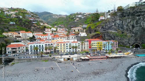 Aerial view of Ponta do Sol, Madeira. Colorful buildings cascade down hillsides to a pebble beach under an overcast sky, capturing the town's unique charm.