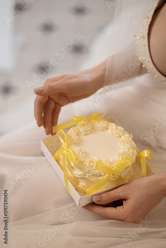 Caucasian Bride Holding Decorative Mini Cake On Lap With Pearl Embellished Dress And Tulle, Delicate Yellow Bows And Ribbon Suggesting Wedding Keepsake And Gentle Preparation Scene