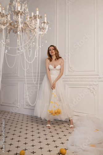 Bride In Tulle Gown Holding Lemons, Caucasian Model Posed Under Chandelier Pearls In Ornate White Studio, Clutching Mesh Bag Of Citrus, Calm Gaze, Editorial Bridal Campaign Vibe With Soft