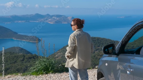 Woman on a road trip admiring beautiful sea view. Female traveler standing near her car on a mountain road, enjoying the stunning panoramic view of the blue sea and islands during a scenic journey