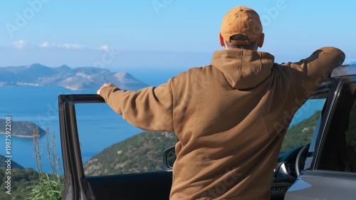Man enjoying a beautiful sea view during a car road trip. Relaxed man wearing a hoodie and a cap enjoying a scenic view of the sea and islands during a road trip, leaning on his car door with his arms