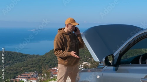 Frustrated man calling for help with a broken down car on a road trip. Stranded man on a scenic roadside, anxiously talking on his phone for assistance next to his car with an open hood