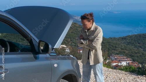 Stressed woman waiting for roadside assistance. Frustrated woman standing next to her broken-down car with the hood open, stranded on a scenic mountain road overlooking the sea and waiting for help
